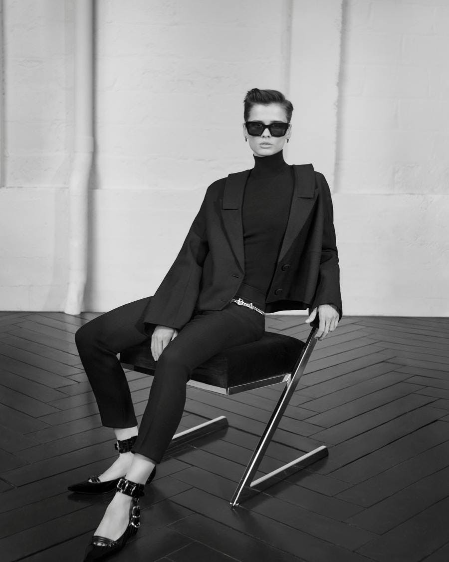 Black and white image of a model in all black with sunglasses sitting on a chair