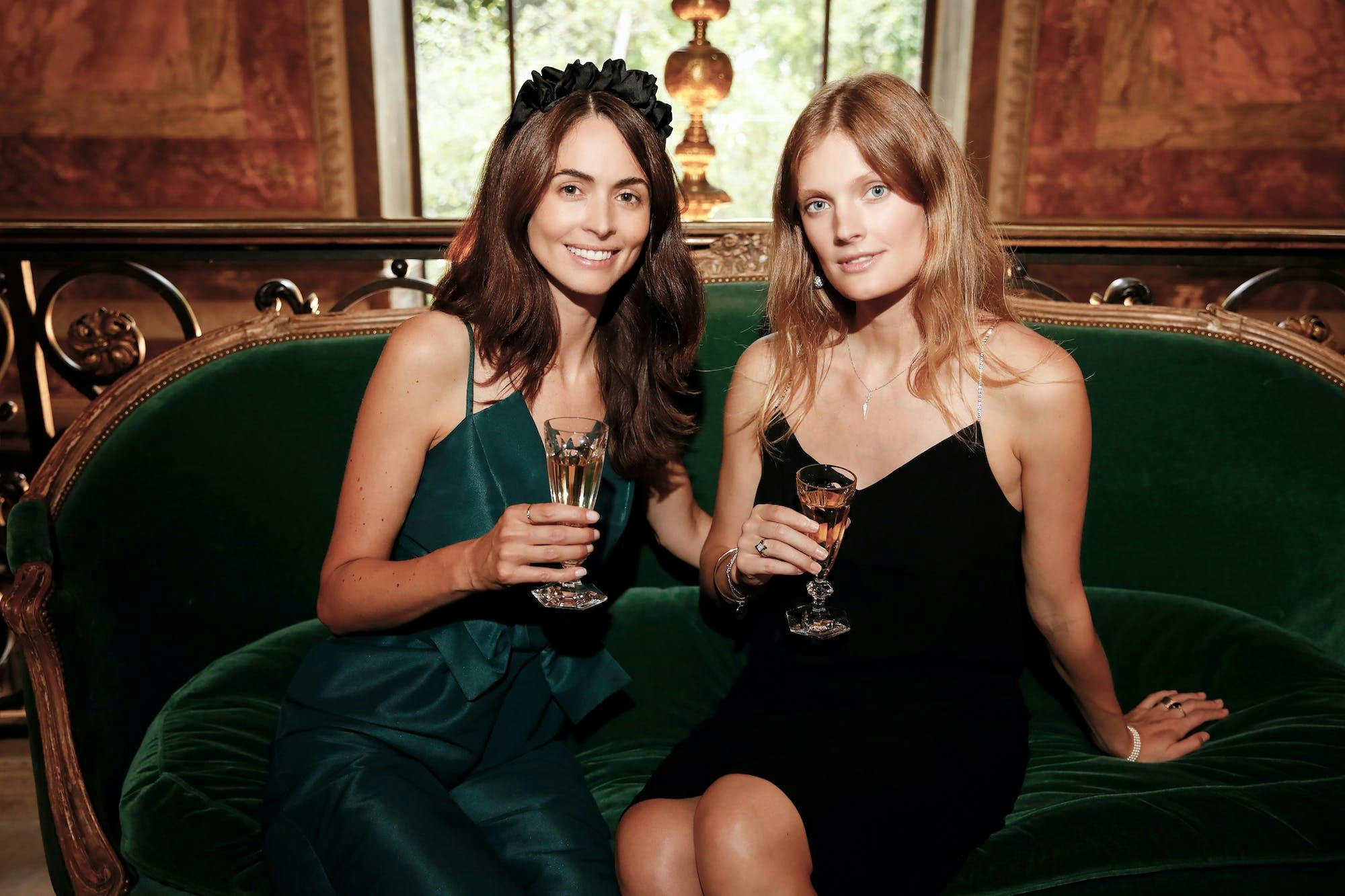 Two women in dresses sitting on a coach holding champagne glasses