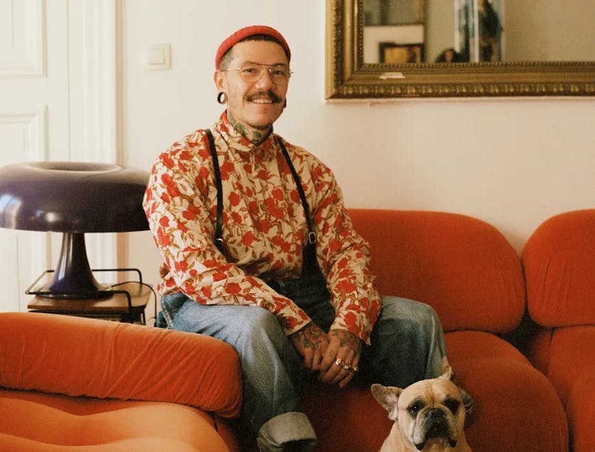 Adrian Appiolaza sitting on a couch with a dog by his feet. He is wearing an orange floral top, jeans, suspenders, brown sneakers and an orange beanie.