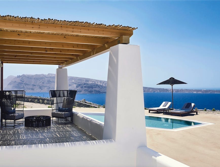 A covered porch with two chairs facing a pool and a beautiful view of the water in Santorini.