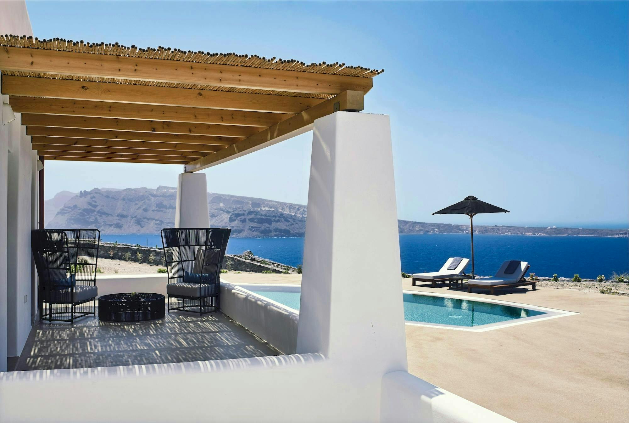 A covered porch with two chairs facing a pool and a beautiful view of the water in Santorini.