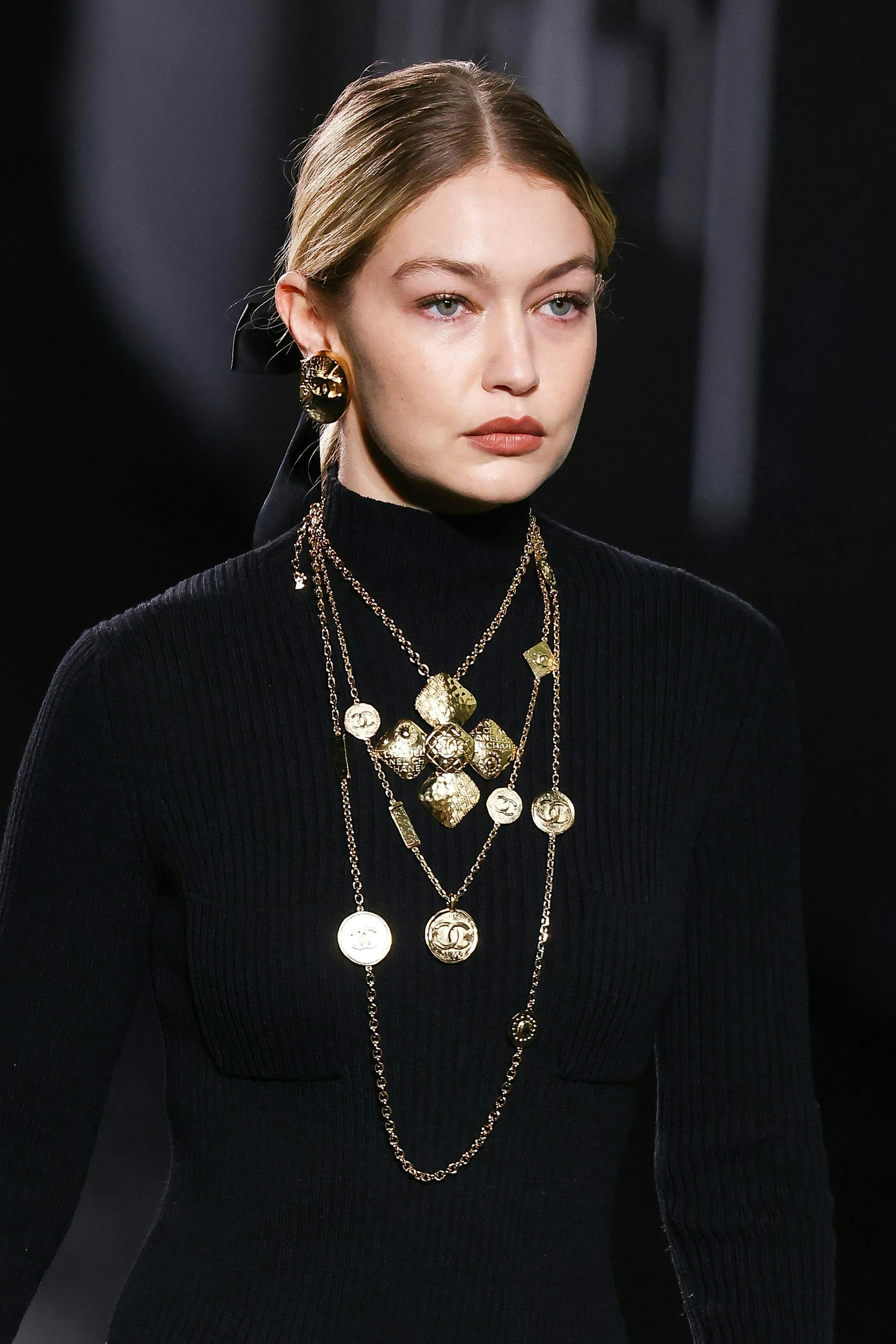 Fall/Winter 2024 Beauty Trends: A closeup of Gigi Hadid walking down the runway for Chanel wearing a black turtleneck and gold necklaces