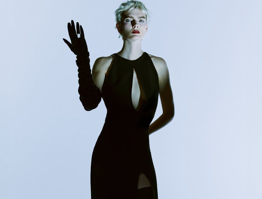 A model with a fitted black dress with a slit at the chest, black elbow-length gloves and a ring on their right hand.